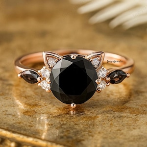 Könnte beinhalten: Roségoldener Ring mit einem großen, runden schwarzen Edelstein in der Mitte, flankiert von kleineren weißen und schwarzen Steinen. Das Ringdesign umfasst katzenohr-förmige Akzente. Der Markenname "jeeyajewels" ist sichtbar.