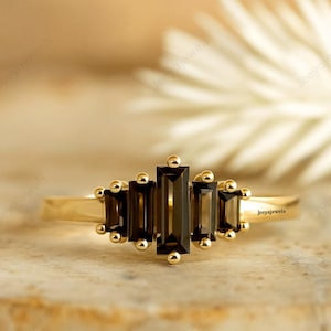 May include: A gold ring with five rectangular, dark brown gemstones. The ring has a delicate band and small gold accents. The ring is displayed on a textured, light brown surface.