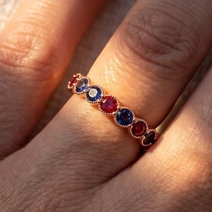 May include: A rose gold band ring featuring a series of alternating round-cut red and blue gemstones set in a bezel style. The ring displays a repeating pattern of vibrant red and deep blue stones secured within a delicate metal frame.