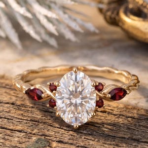 May include: A gold ring featuring a large oval-cut clear centre stone flanked by smaller marquise-cut and round-cut deep red stones. The band has a textured, wavy design, and the setting holds the stones in a prong arrangement on a gold metal base.