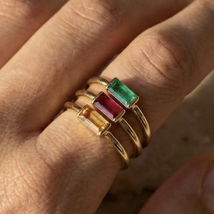 May include: Gold rings with rectangular gemstones in yellow, red, and green are stacked on a finger. The rings have a thin band and a simple design, showcasing the colorful stones. The background is a soft, neutral tone.