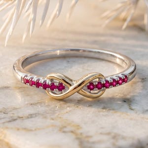 Könnte beinhalten: Ein silberfarbener Ring mit einem goldenen Unendlichkeitssymbol in der Mitte. Der Ring ist mit kleinen, runden, leuchtend roten Steinen besetzt, die entlang der Seiten vom zentralen Unendlichkeitssymbol ausgehen und ein durchgehendes Design bilden.