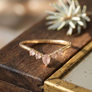 Pode incluir: Um anel de ouro com uma banda em forma de V cravejada com cinco pedras rosa-pálido. A pedra central tem formato de pera, ladeada por duas pedras redondas menores de cada lado, todas presas por garras douradas ao longo da curva superior.