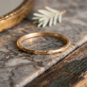 Op de afbeelding: Een gouden bandring met kleine, sprankelende edelstenen gelijkmatig verdeeld over de band. De ring wordt getoond op een marmeren oppervlak met een takje groen en een goudkleurige spiegel op de achtergrond.