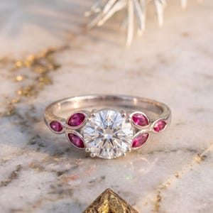 Pode incluir: Um anel de tom prateado com uma grande pedra central redonda e transparente, ladeada por seis pedras mais pequenas de cor rosa intenso com lapidação marquise ao longo da banda. O anel tem um acabamento metálico polido e cravação.