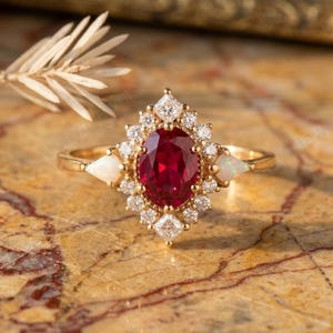 Könnte beinhalten: Ein goldener Ring mit einem zentralen oval geschliffenen roten Edelstein, umgeben von kleineren Diamanten und weißen Edelsteinen. Der Ring hat ein Halo-Design. Zwei weiße Edelsteine flankieren den Hauptstein. Das Design ist klassisch und elegant.