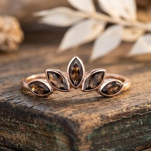 May include: A rose gold ring featuring five marquise-cut smoky quartz gemstones arranged in a fan-like crown shape. The band is smooth and polished, holding the dark brown, translucent stones in individual bezel settings that follow a curved arc.