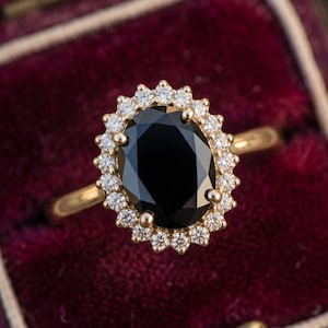 Puede incluir: Un anillo de oro con una gran piedra central negra de corte ovalado, rodeada por un halo de pequeñas piedras blancas redondas y transparentes. El anillo tiene una banda de oro pulido y un engaste de garras con un diseño clásico.