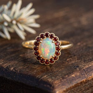 May include: A gold ring featuring a central oval-shaped iridescent opal surrounded by a halo of small, round, deep red garnet stones. The setting has a scalloped, beaded edge, and the band is a smooth, polished gold metal finish.