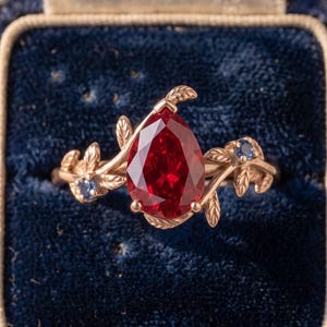 Könnte beinhalten: Ein Ring aus Roségold mit einem großen, birnenförmig geschliffenen roten Edelstein als Mittelpunkt. Die Ringschiene ist mit detaillierten Metallblättern und Ranken verziert, ergänzt durch zwei kleine runde blaue Steine im Blattwerk.