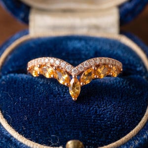 Peut inclure: Une bague en forme de V en or rose, ornée d'une rangée de citrines jaunes taille marquise sur le bord inférieur et d'une ligne de petits diamants ronds et transparents sertis pavé le long de la courbe supérieure de l'anneau.