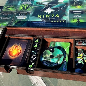 May include: A wooden box with compartments for storing game pieces. The box has a black interior and is labelled "Ninja: Assassinate!" The box contains a green and black card with a ninja figure, a red and orange card with a fire symbol, a stack of green dice, and a stack of black and green tokens.