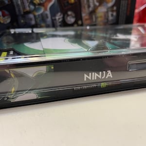 May include: A black and green card game box with the word "NINJA" printed on the front. The box has a clear plastic cover and the text "HERO COMPLEXITY" is printed below the word "NINJA".