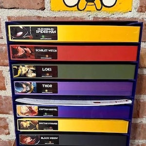 Peut inclure: Unité de rangement bleue à plusieurs niveaux, remplie de boîtes colorées portant les noms de personnages Marvel tels que Spider-Man, Scarlet Witch et Thor. Une brique jaune peinte avec un visage de dessin animé est au-dessus. Les boîtes sont de couleurs variées.