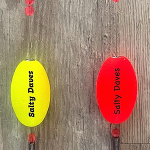 May include: Two fishing floats, one yellow and one red, suspended by clear line. Each float has the text "Salty Daves" printed on it. The floats are attached to a series of red beads and a small weight.