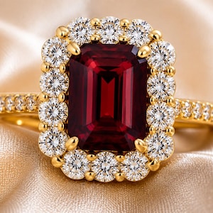 Puede incluir: Un anillo de oro con una gran piedra preciosa de color rojo intenso y corte esmeralda rectangular en el centro. La piedra central está rodeada por un halo de pequeños diamantes blancos redondos de talla brillante, con diamantes adicionales en la banda.