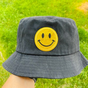 May include: Black bucket hat with a yellow smiley face patch. The hat has a wide brim and is made of a soft, comfortable fabric.