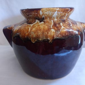 Könnte beinhalten: Eine Keramikvase mit breitem, rundem Körper und zwei seitlichen Griffen. Die Vase hat einen dunkelbraunen Boden, der in ein helleres braunes und weiß gesprenkeltes Oberteil übergeht. Der Innenraum ist dunkelblau.