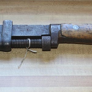 Puede incluir: Una llave ajustable con una cabeza de metal oscuro y un mango de madera. La llave está colocada horizontalmente sobre una superficie de madera de color marrón claro. El mango está sujeto a la cabeza de metal con remaches.