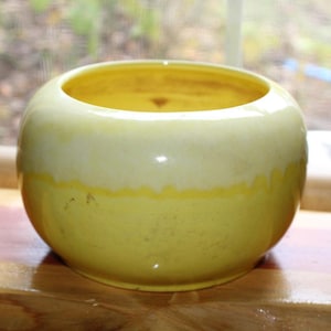 May include: A round, ceramic bowl with a glossy finish. The bowl is primarily yellow, with a lighter, creamy white band around the top. The interior is also yellow. The bowl sits on a wooden surface.
