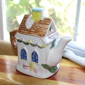 May include: A white ceramic teapot shaped like a house with a brown roof, blue arched windows, and a chimney. The teapot has a white spout and handle, and is decorated with green garland and pink and yellow accents.