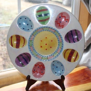 May include: A white ceramic Easter egg serving platter with a central yellow section and eight egg-shaped indentations. Each indentation is painted with a different colorful egg design, including stripes, dots, and zigzags. The platter is displayed on a wooden stand.