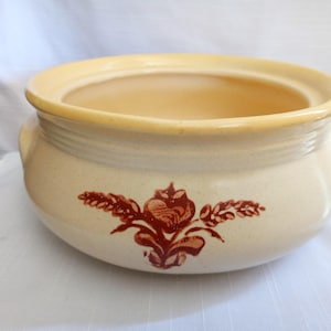 May include: A cream-coloured ceramic bowl with a yellow rim and a floral design in shades of brown. The bowl has a rounded shape with two small handles and a lid. This vintage-style bowl is suitable for serving food.