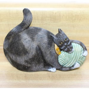May include: A grey and white tabby cat ornament with a green ball of yarn. The cat is lying down, with its head resting on the yarn. The cat has yellow eyes and a gold collar. The ornament is on a light brown surface.