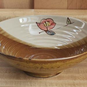 May include: A ceramic serving bowl with a floral design. The bowl has a wide, shallow shape with a brown exterior and a cream-colored interior. A red and yellow flower with green leaves is painted on the inside.