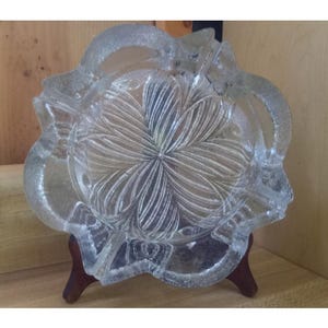 Puede incluir: Cenicero de vidrio transparente con diseño floral. El cenicero tiene un borde festoneado y está sostenido por un pequeño soporte de madera. El patrón floral está grabado en el vidrio, creando una superficie texturizada.