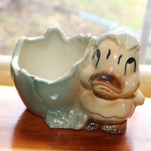 May include: A ceramic planter shaped like a duckling hatching from an egg. The planter is primarily beige with green and brown accents. The duckling has black eyes and a brown beak. The eggshell is light green and beige.
