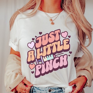 May include: White t-shirt with a pink, orange, and yellow graphic that says "Just a Little Pinch" with a cartoon syringe and hearts.
