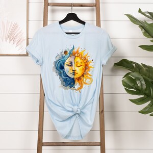 May include: A light blue t-shirt with a watercolor design of a sun and moon with faces. The sun is yellow and orange, and the moon is blue and white.