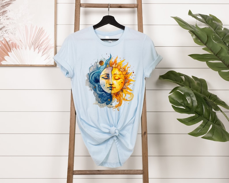 May include: A light blue t-shirt with a watercolor design of a sun and moon with faces. The sun is yellow and orange, and the moon is blue and white.