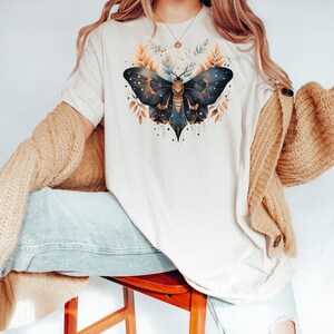 May include: A white t-shirt with a gold and blue moth illustration. The moth has intricate details and is surrounded by gold leaves.