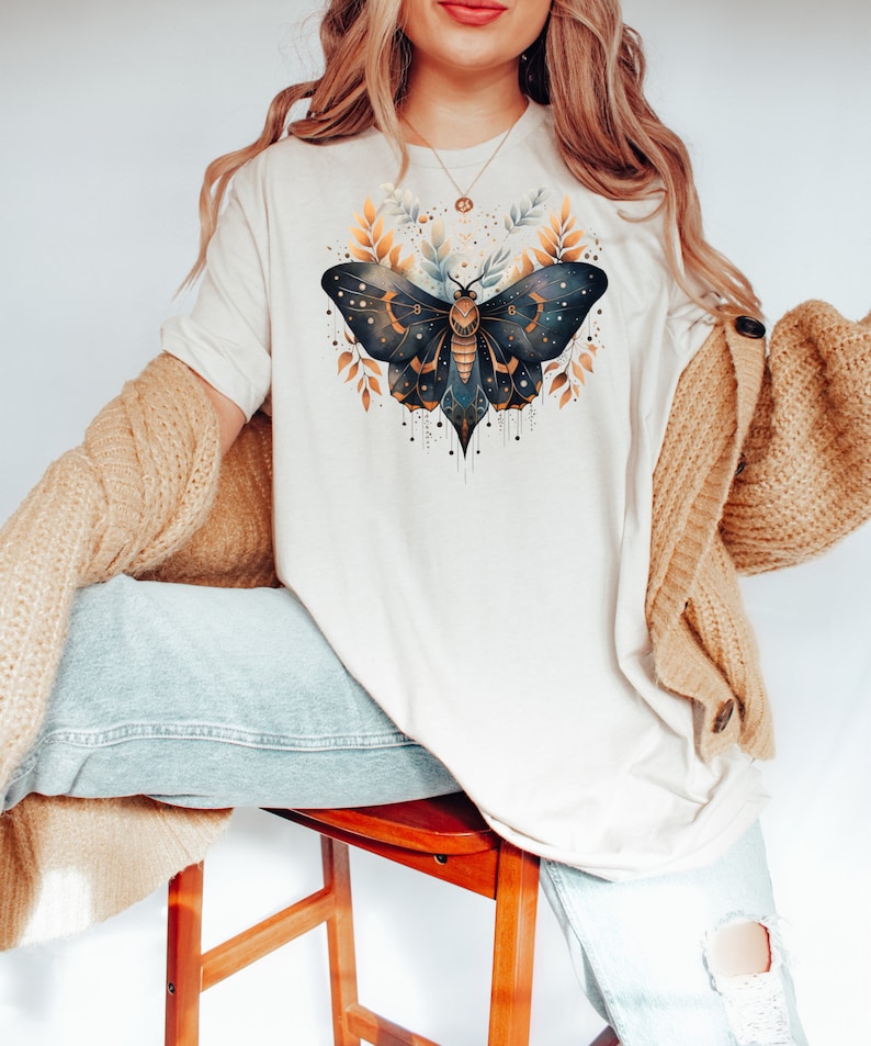 May include: A white t-shirt with a gold and blue moth illustration. The moth has intricate details and is surrounded by gold leaves.