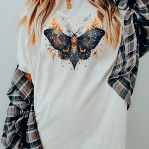 May include: A white oversized t-shirt with a graphic of a black and gold butterfly with intricate details. The butterfly is surrounded by gold leaves and has a celestial design.