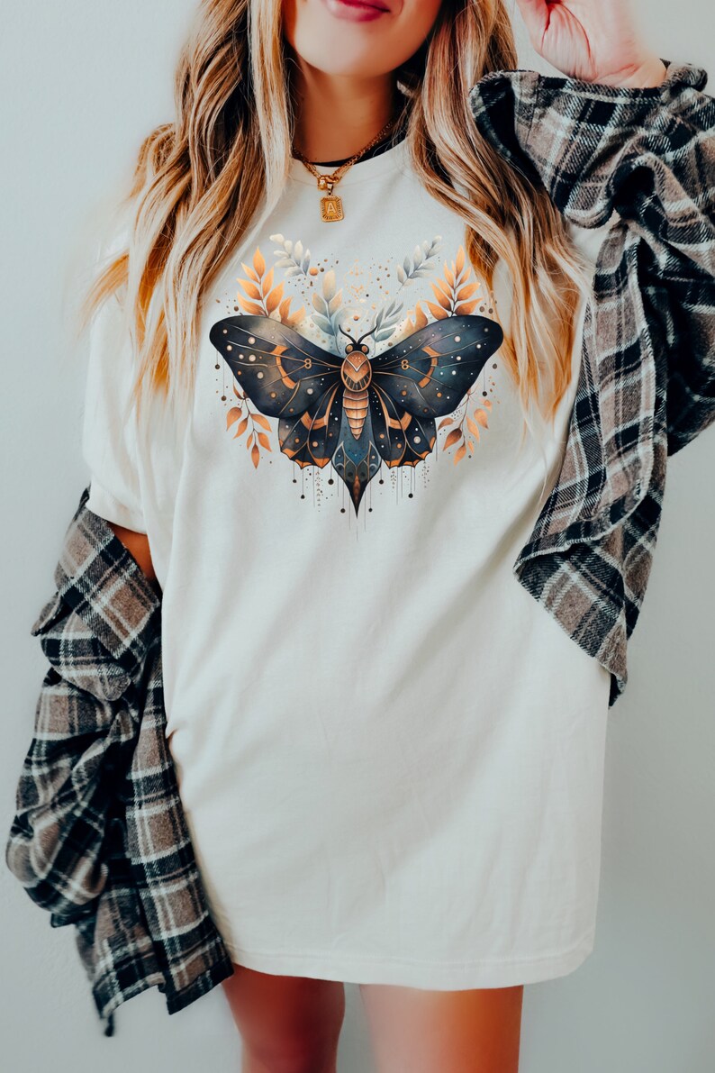 May include: A white oversized t-shirt with a graphic of a black and gold butterfly with intricate details. The butterfly is surrounded by gold leaves and has a celestial design.