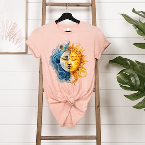 May include: A peach-colored t-shirt with a watercolor design of a sun and moon with faces. The sun is yellow and the moon is blue.