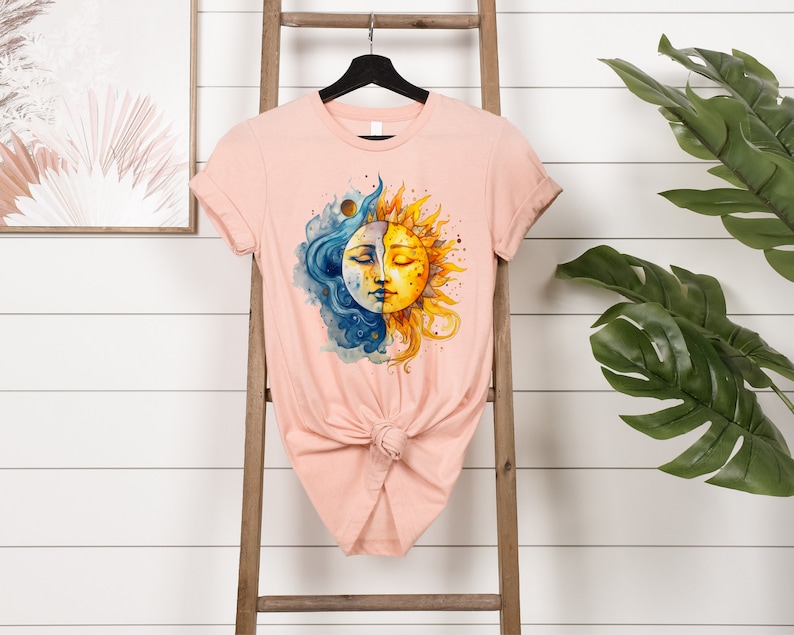 May include: A peach-colored t-shirt with a watercolor design of a sun and moon with faces. The sun is yellow and the moon is blue.