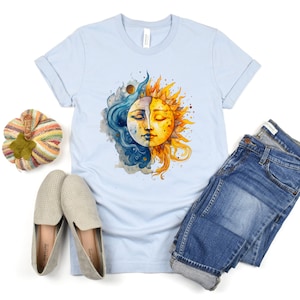May include: A light blue t-shirt with a watercolor illustration of a sun and moon. The sun is yellow and orange with a face, and the moon is blue and white with a face. The sun and moon are overlapping.