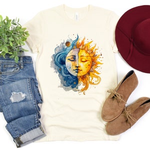 May include: A cream colored t-shirt with a watercolor illustration of a sun and moon with faces. The sun is yellow and the moon is blue. The design is centered on the shirt.
