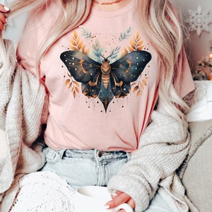 May include: A pink t-shirt with a black and gold butterfly graphic. The butterfly has intricate details and is surrounded by gold leaves.