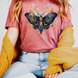 May include: A woman wearing a pink t-shirt with a dark blue and gold butterfly graphic. The butterfly has a celestial design with stars and moons. The woman is also wearing blue jeans and a yellow cardigan.