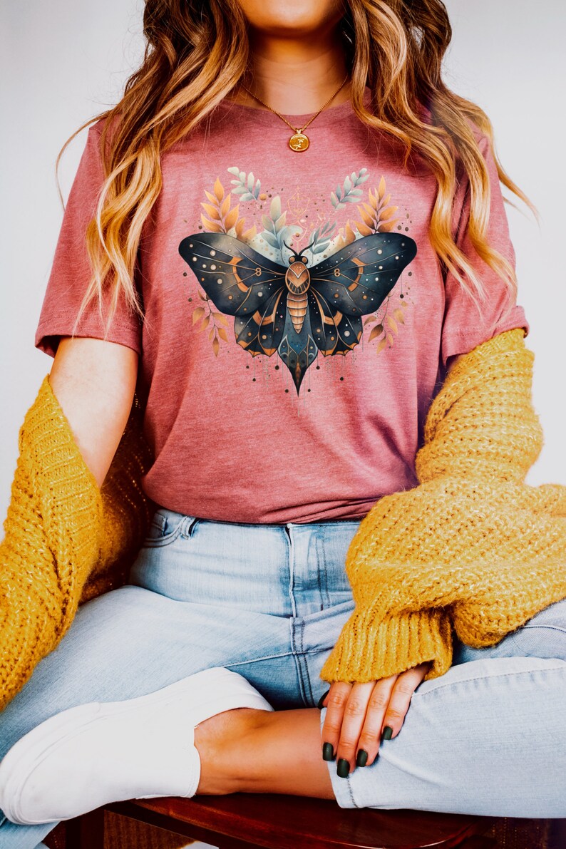 May include: A woman wearing a pink t-shirt with a dark blue and gold butterfly graphic. The butterfly has a celestial design with stars and moons. The woman is also wearing blue jeans and a yellow cardigan.