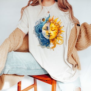 May include: A white t-shirt with a watercolor illustration of a sun and moon with faces. The sun is yellow and the moon is blue.
