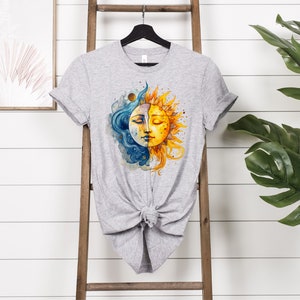 May include: A gray t-shirt with a watercolor design of a sun and moon. The sun is yellow and orange with a smiling face. The moon is blue and white with a face that is half closed eyes. The design is centered on the shirt.