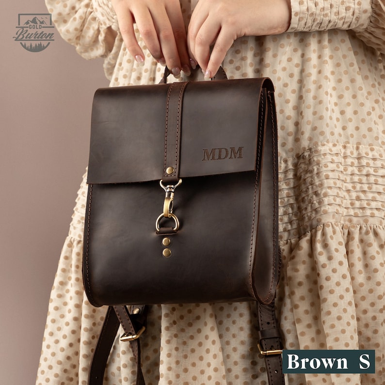 May include: A dark brown leather backpack with a flap closure and adjustable straps. The backpack features gold-colored hardware and the initials "MDM" engraved on the front. The bag is a small size.