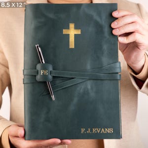 May include: A dark green leather journal with a gold cross and the initials "F.J. EVANS" embossed on the front. The journal has a leather strap with a gold buckle and a pen tucked into the strap.