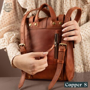 May include: A copper-colored leather backpack with adjustable straps, a top handle, and a front zipper. The backpack has brass-colored hardware and is labeled "Copper S". The "Gold Burton" logo is visible in the upper left corner.